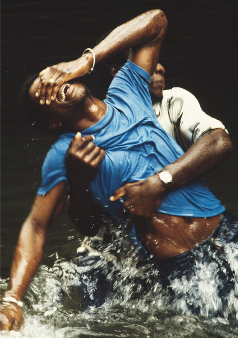 Zwei Männer in T-Shirt und Jeans stehen bis zur Hüfte im Wasser, der hintere hält den vorderen, sich in Trance windenden an der Hüfteumfasst