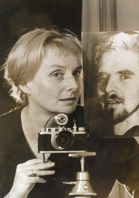 Sepia-Porträt einer blonden Frau mit älterer Kamera auf Tischstativ neben  lebensgroßem Fotoporträt eines vollbärtigen Mannes