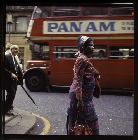 Eine Frau geht über die Straße, hinter ihr ein roter Doppeldeckerbus mit der Werbeaufschrift "Pan Am"