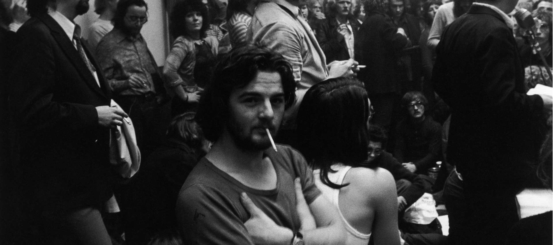 Viele Menschen in einem Raum bei einer Versammlung, ein junger Mann mit Zigarette schaut direkt in die Kamera