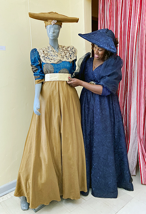 Eine Frau mit langem Kleid und horizontal sehr ausladendem Hut steht stolz neben einer Modepuppe in ähnlicher Bekleidung