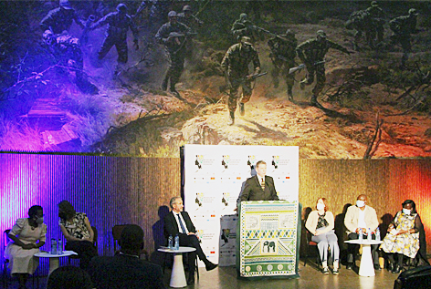 Redner auf einem teppichbehängtem Podium vor überdimensioniertem Wandbild, das bewaffnete Soldaten zeigt, links und rechts neben ihm sitzend jeweils drei weitere Personen