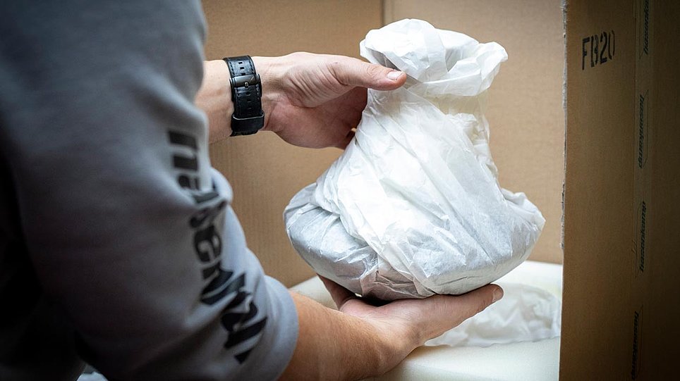 Hände packen ein in Papier eingepacktes Objekt in eine Kiste mit Schaumstoffauskleidung (öffnet Vergrößerung des Bildes)