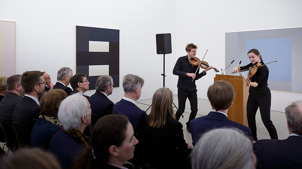 Zwei stehende Violinistinnen spielen vor sitzendem Publikum (öffnet Vergrößerung des Bildes)