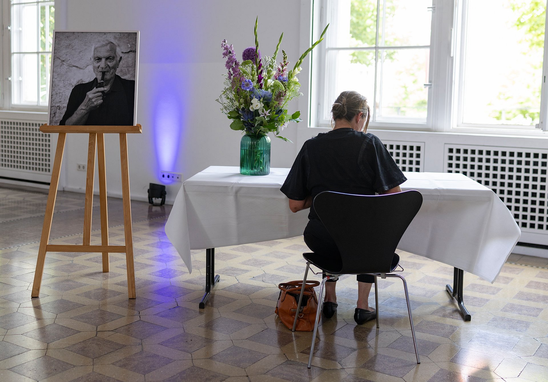 Eine Frau sitzt mit dem Rücken zum Fotografierenden auf einem Stuhl und schreibt in ein Buch, links neben ihr ein großformatiges Schwarz-Weiß-Porträtfoto eines Mannes