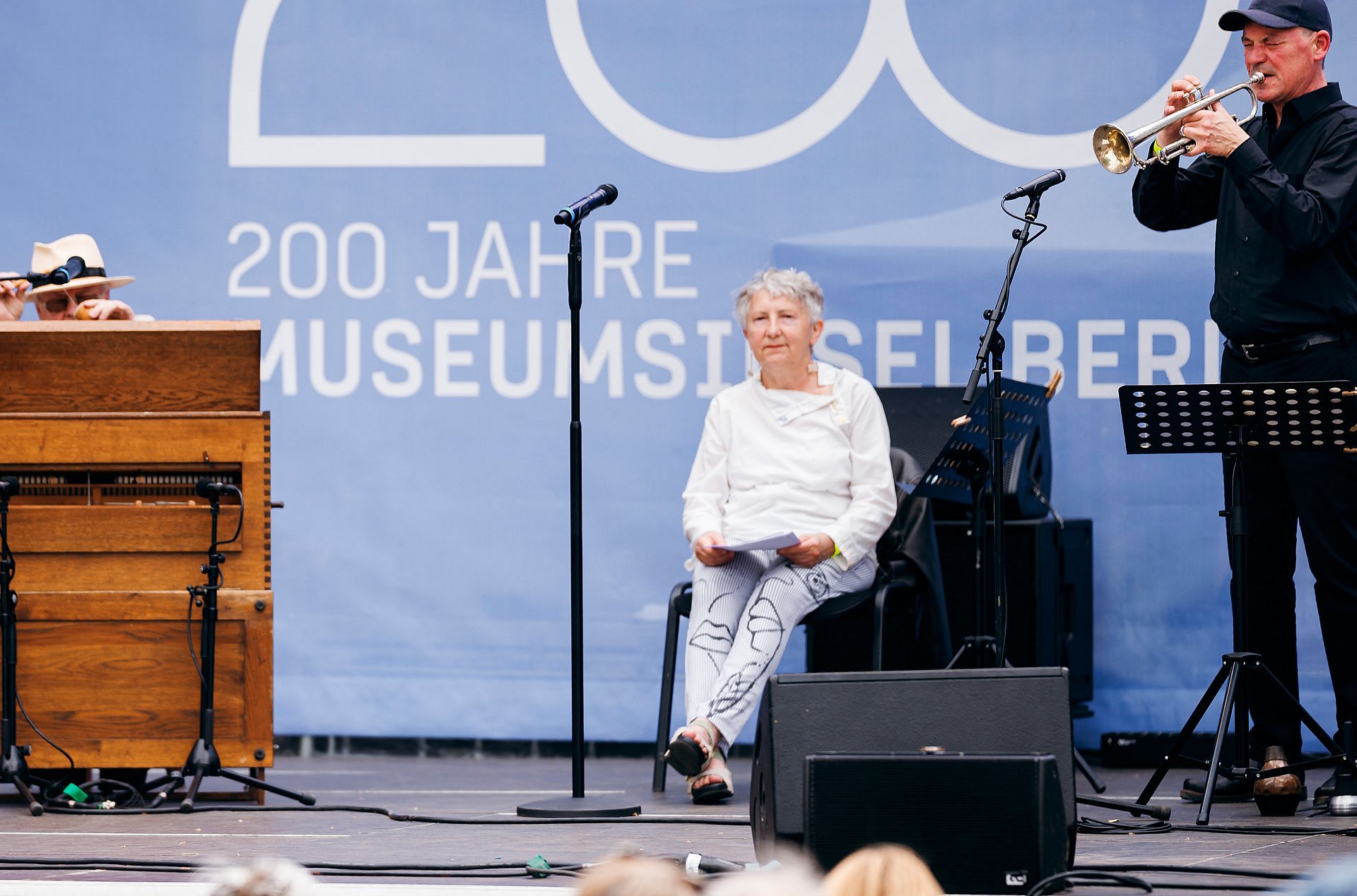Eine Frau sitzt auf einem Stuhl auf einer Bühne, rechts spielt ein Mann Trompete