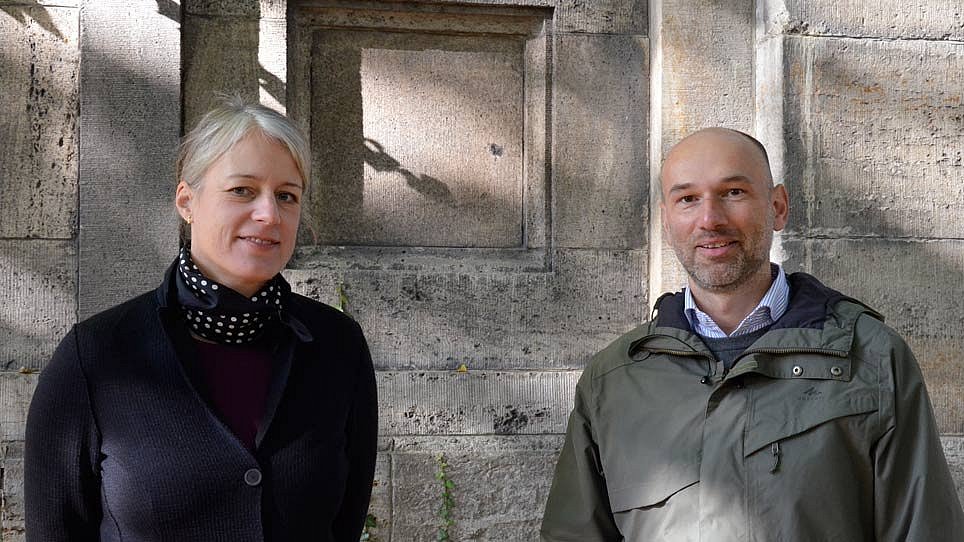 Eine Frau und ein Mann stehend lächelnd vor einer grauen Mauer