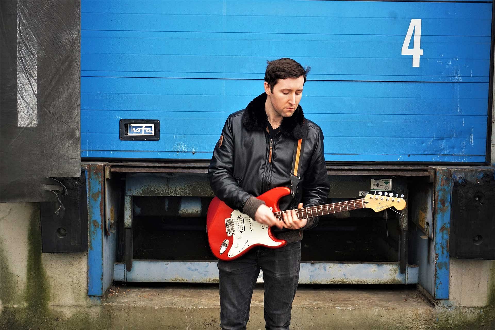 Mann steht mit einer roten E-Gitarre vor einem blauen Gebäude