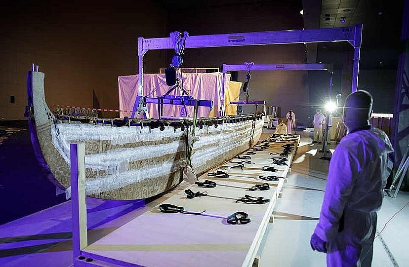 Links im Bild befindet sich ein hölzernes Boot, das in einer Haltekonstruktion hängt und auf einer Kiste platziert wird, vorne rechts sieht man einen Mann in weißer Schutzkleidung (öffnet Vergrößerung des Bildes)