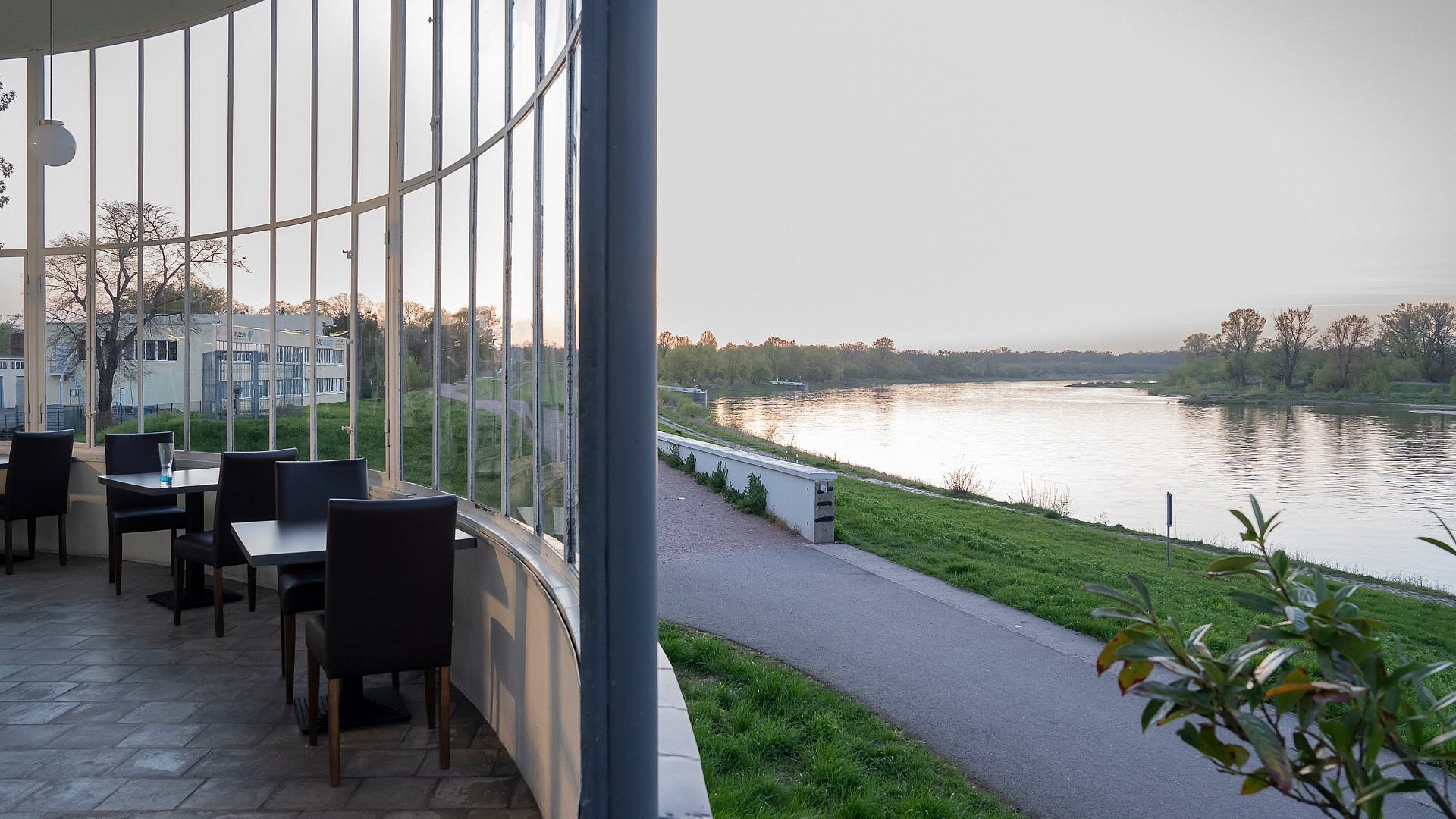 Links im Bild eine verglaste Veranda, rechts ein breiter Fluß mit Uferradweg