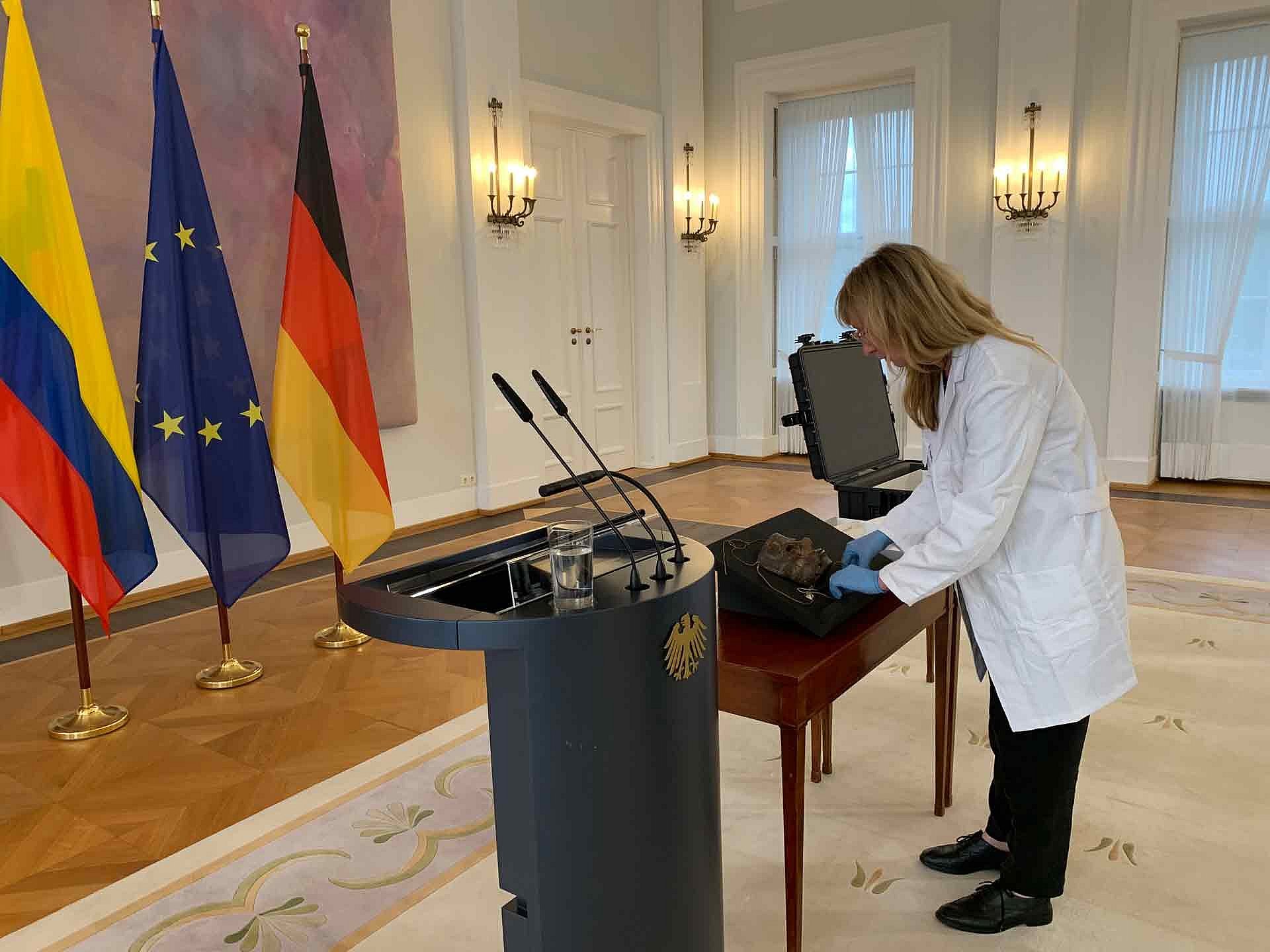 Eine Frau steht in einem repräsentativen Saal vor Flaggen, vor ihr ein Tisch mit zwei Holzmasken