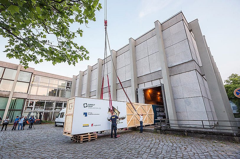 Eine große Holzkiste wird aus einem Gebäude durch ein Rolltor nach draußen geschoben und mit Spanngurten an einem Kran befestigt, während mehrere Personen im Hintergrund das Spektakel beobachten (öffnet Vergrößerung des Bildes)