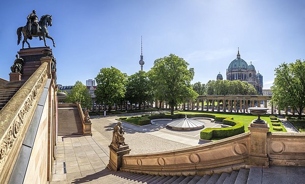 Panoramablick auf die Museumsinsel von der Alten Nationalgalerie aus, man sieht einen Garten und einen Kolonnadengang, die Sonne scheint