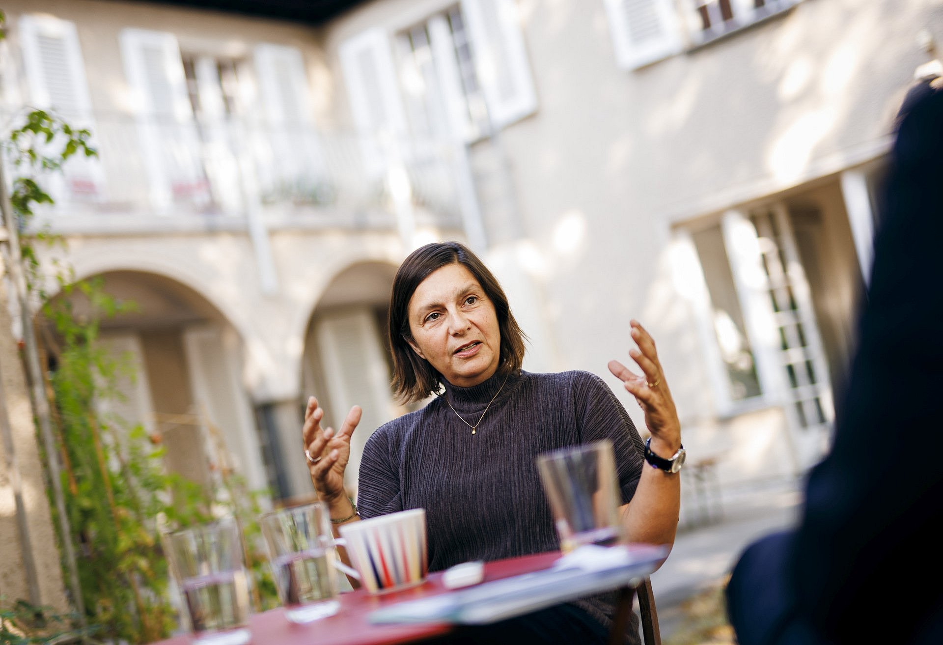 Frau sitzend an einem Tisch, im Gespräch mit den Händen gestikulierend.