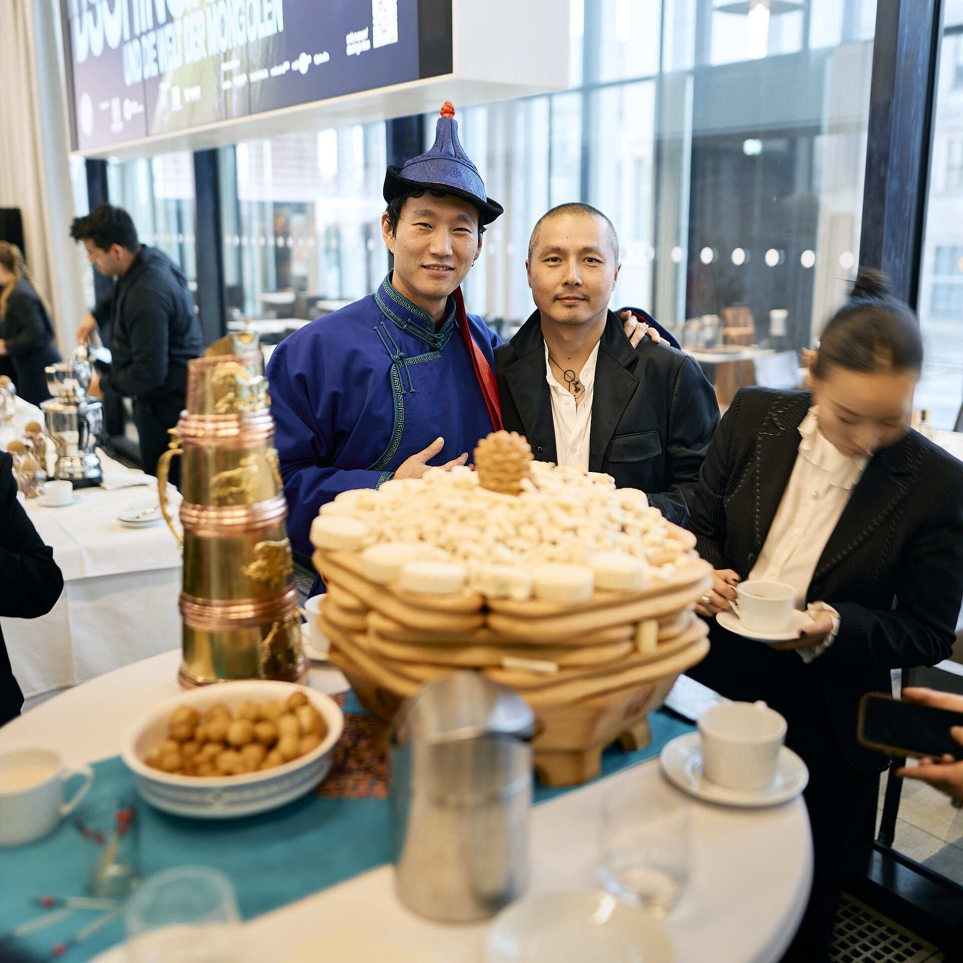 Zwei Männer posieren hinter einer aufgeschichteten Gebäckplatte. Daneben hält eine Frau eine Tasse mit Untertasse.