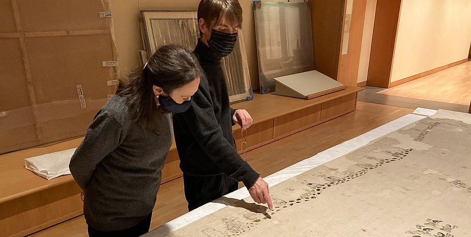 Zwei Personen betrachten ein historisches Baumwolltuch, das auf einem großen Tisch ausgebreitet ist
