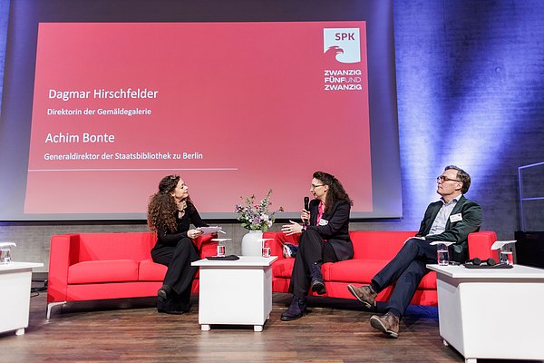 JE25 Drei Personen auf roter Couch auf Bühne