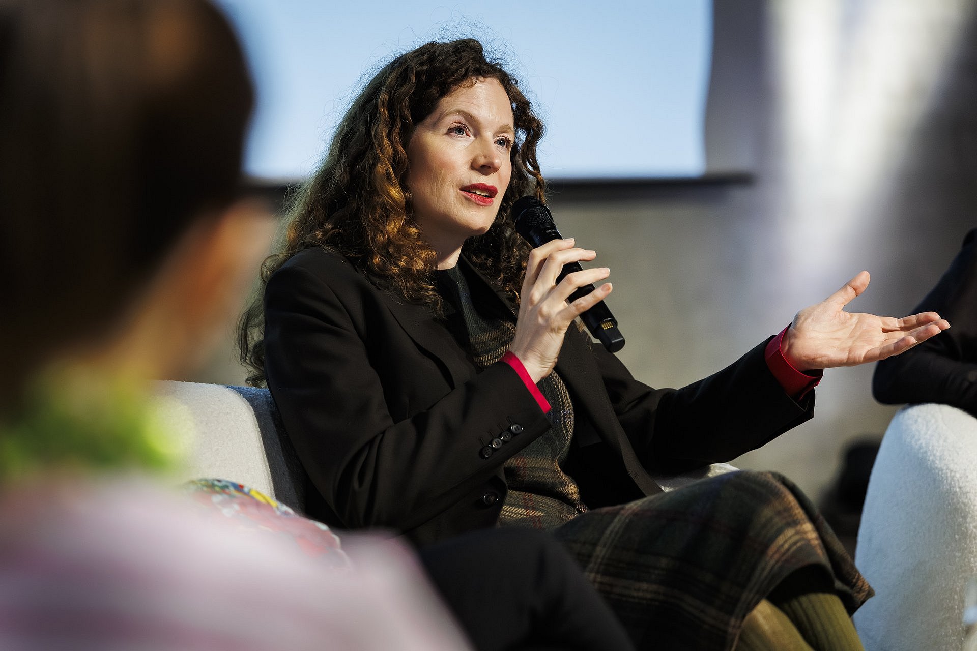 Sandra Richter sitzt auf einem Podium und spricht in ein Mikrofon, während sie mit der Hand gestikuliert.