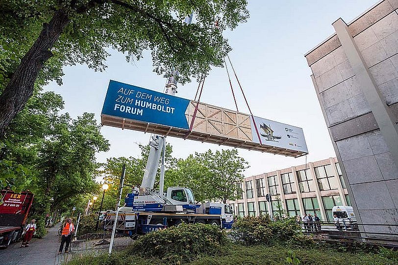 Eine große Holzkiste wird aus einem Gebäude durch ein Rolltor nach draußen geschoben und mit Spanngurten an einem Kran befestigt, während mehrere Personen im Hintergrund das Spektakel beobachten (öffnet Vergrößerung des Bildes)