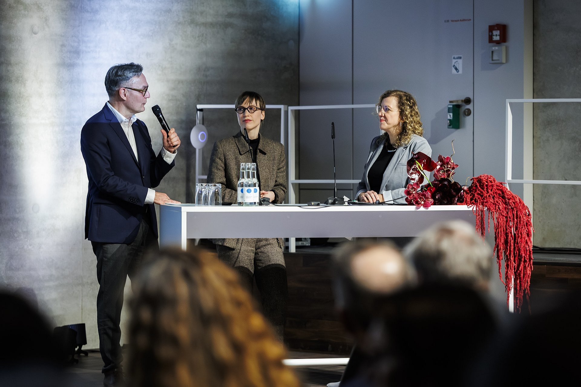 Achim Bonte sitzt auf einem Podium und spricht in ein Mikrofon; neben ihm sitzen zwei Gesprächspartnerinnen bei einer Veranstaltungssituation.