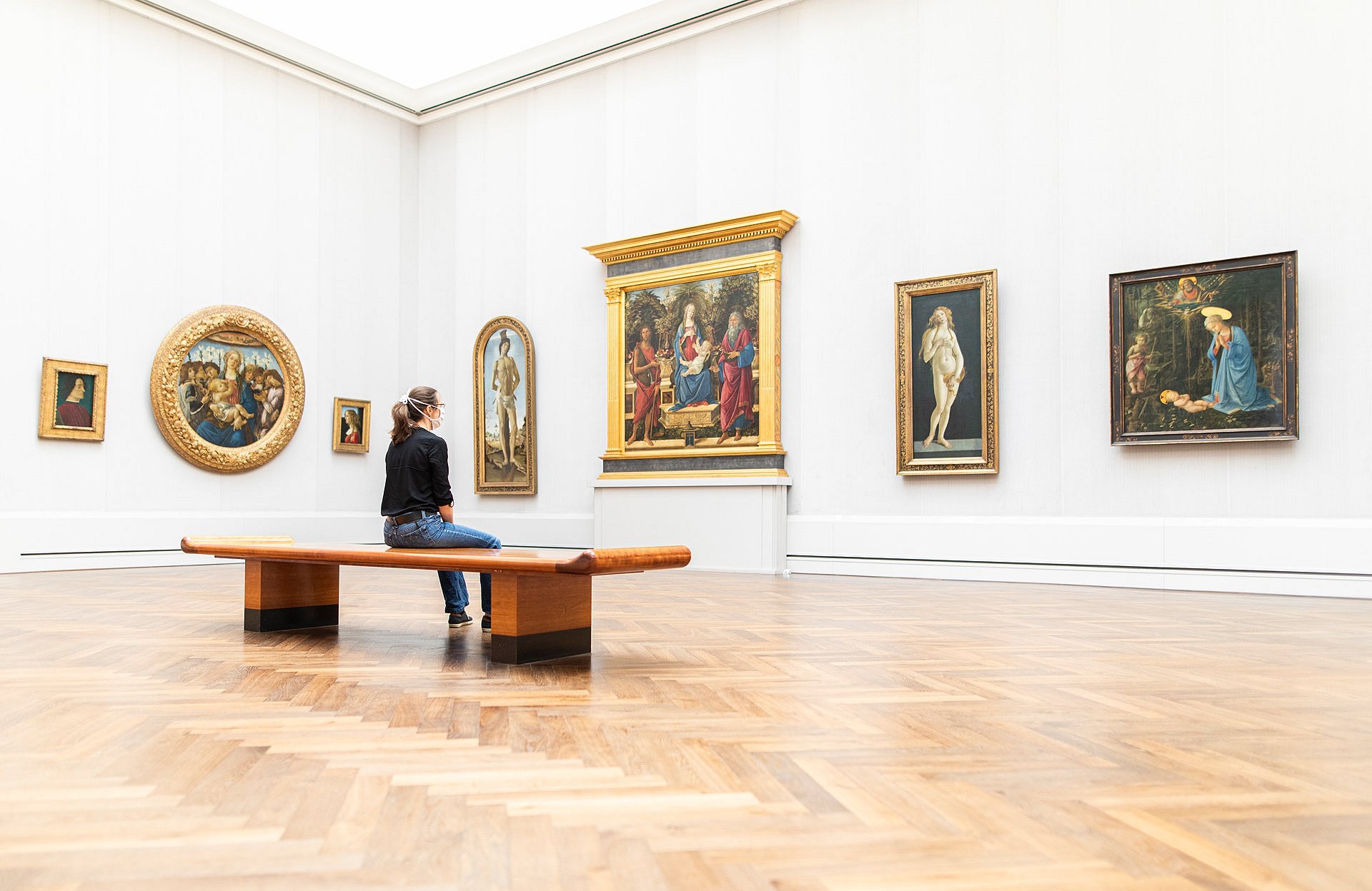 Person mit Mundschutz sitzt allein auf einer Bank in einem Museumssaal mit alten Gemälden