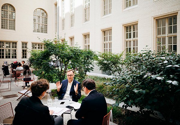 Im Innenhof des Café Felix in der Stabi unter den Linden ging die SPK-Sommerinterviewreihe in die nächste Runde Drei Personen sitzend im Gespräch an einem Tisch in einem Hinterhof