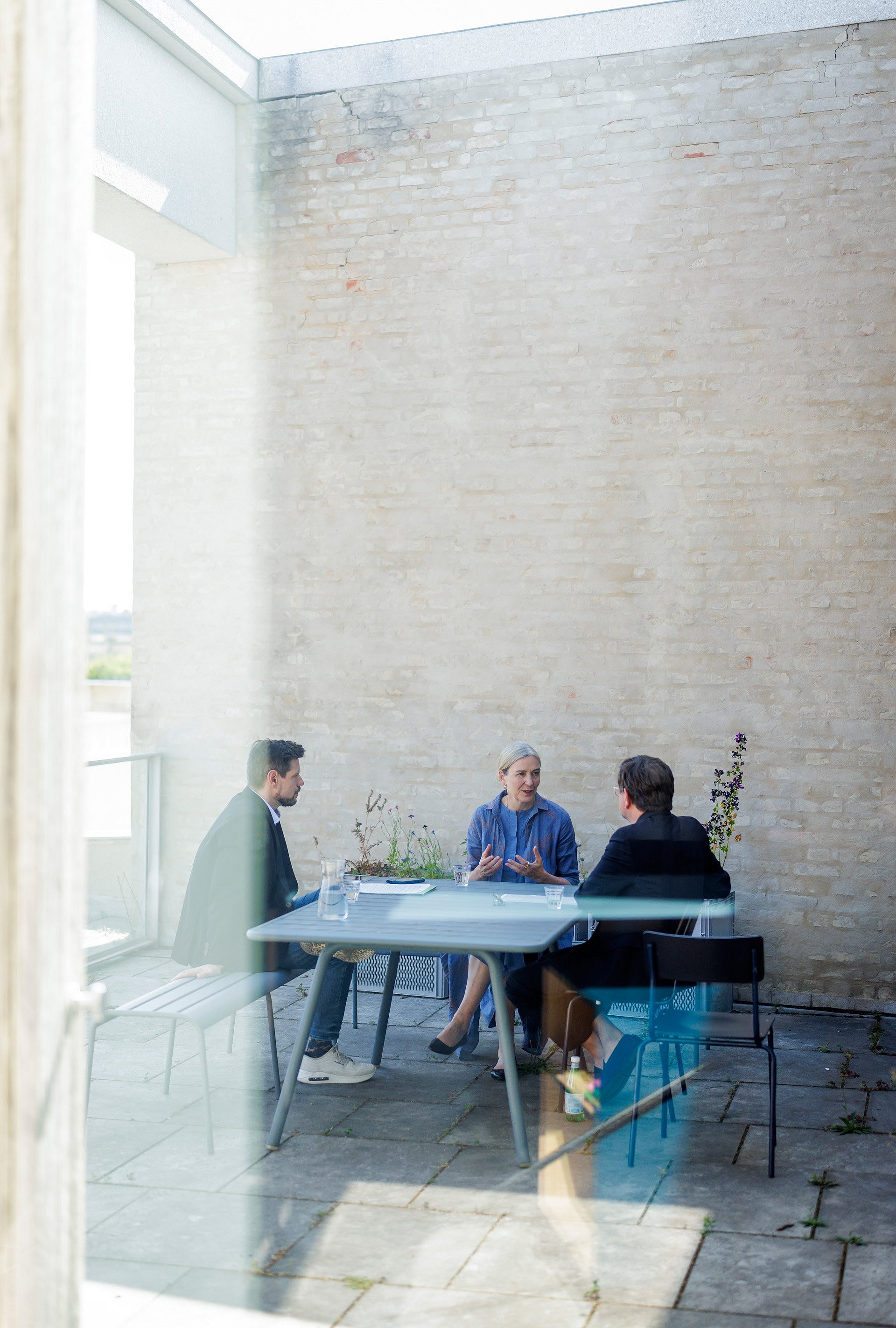 Drei Personen sitzen an einem Tisch auf einer Terrasse vor einer Steinmauer im Gespräch