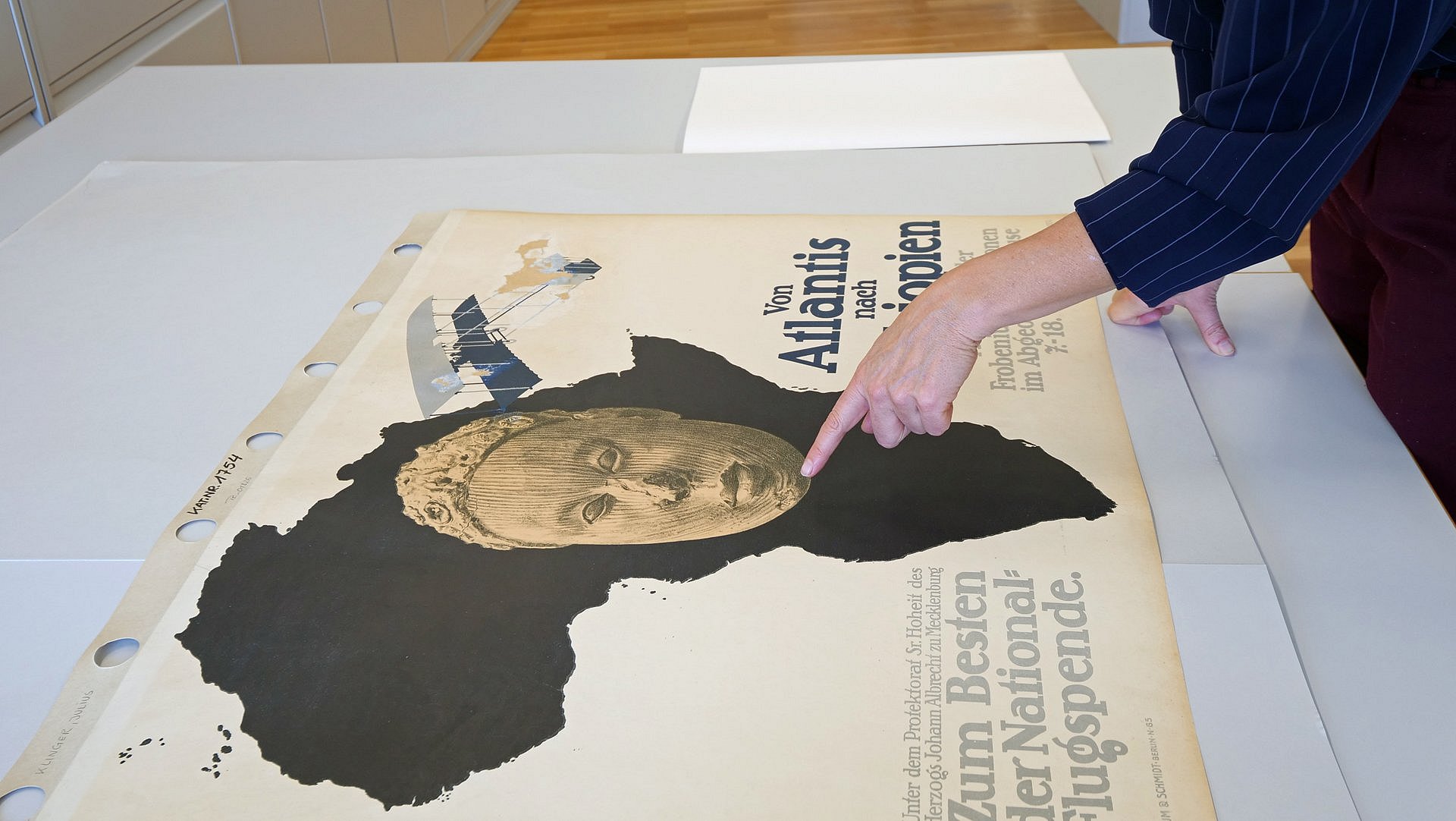 Ein Finger zeigt auf ein Plakat mit dem Umriss Afrikas als monochrom-schwarzer Fläche und der Zeichung eines Kunstobjekts in Form eines Kopfes