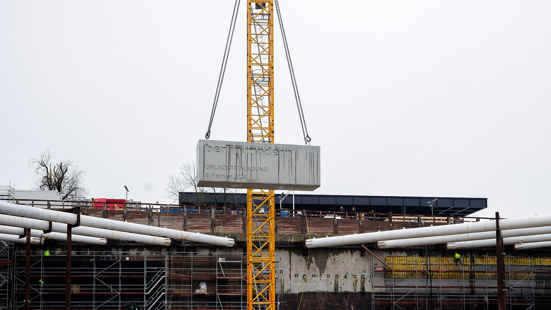 Ein großer Betonquader hängt von einem Kran gehalten über einer Baugrube