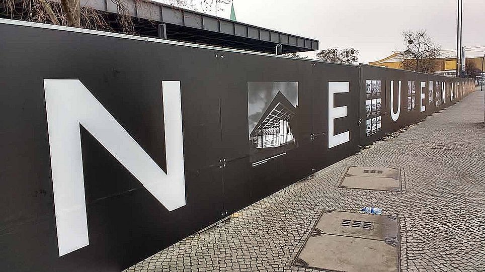 Ein mit Plakaten beklebter, schwarzer Baustellenzaun zieht sich durch das Bild (öffnet Vergrößerung des Bildes)