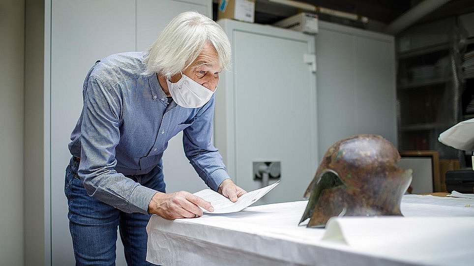 Ein Mann mit einem Blatt Papier studiert ein vor ihm liegendes Metallobjekt in Form eines Kriegerhelms (öffnet Vergrößerung des Bildes)