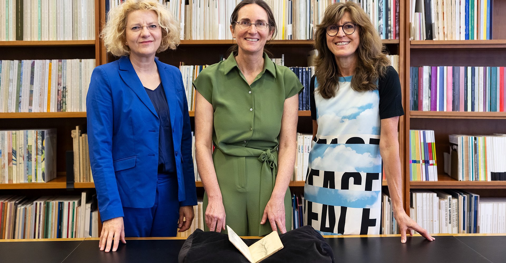 Drei Frauen stehen vor einem Bücherregal und präsentieren ein Notizbuch