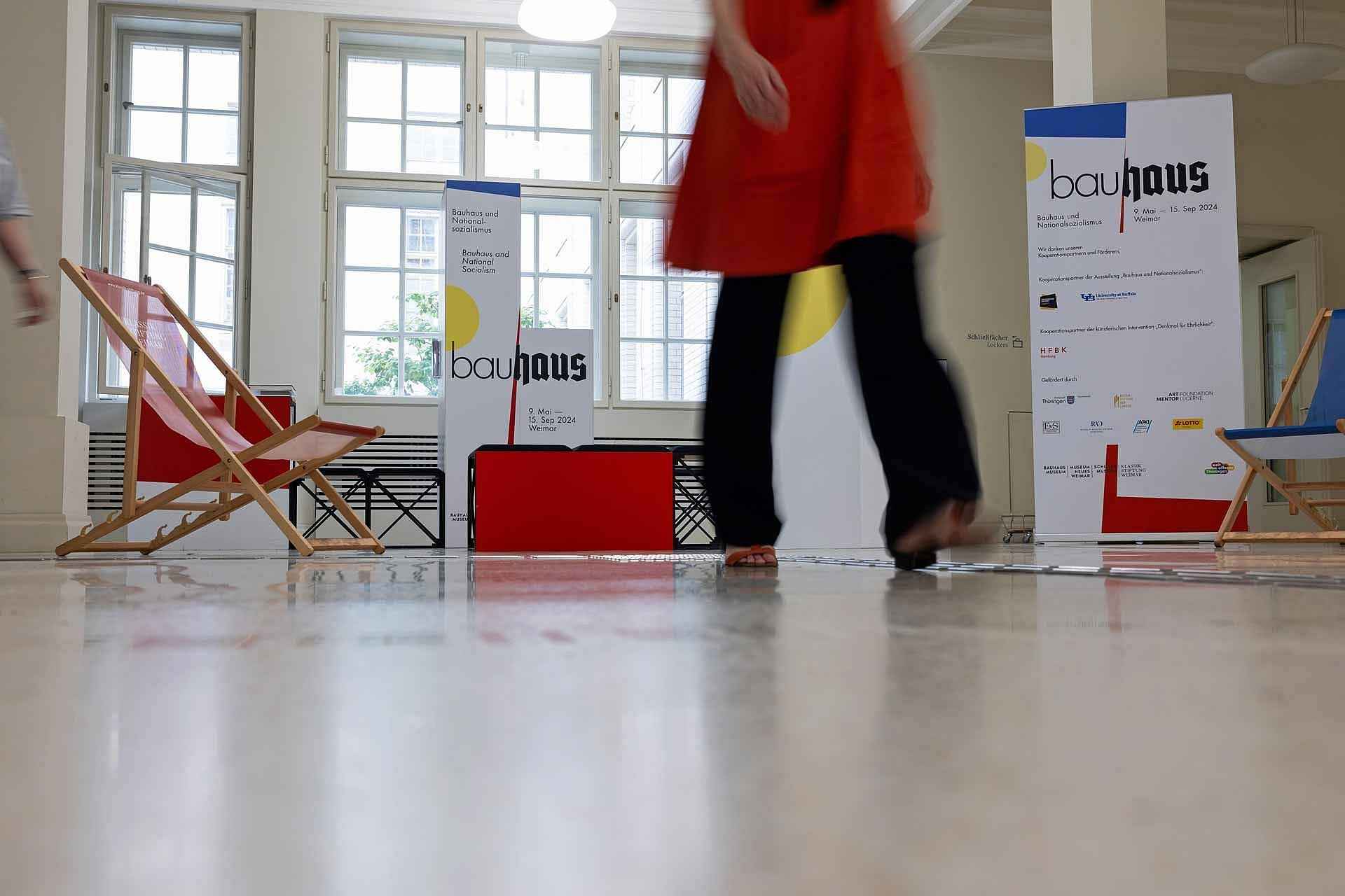 Liegestühle und beschriftete Aufsteher in einem Foyer, im Vordergrund verschwommen Unterteil einer gehenden Person