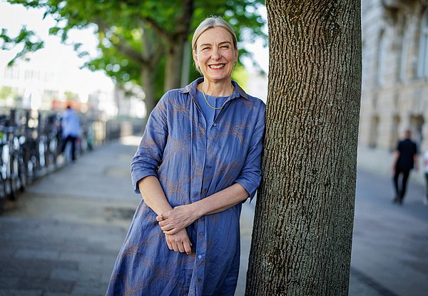 Seit 1. Juni 2025 die neue SPK-Präsidentin Eine Frau lehnt in einer sommerlichen urbanen Umgebung an einem Baum