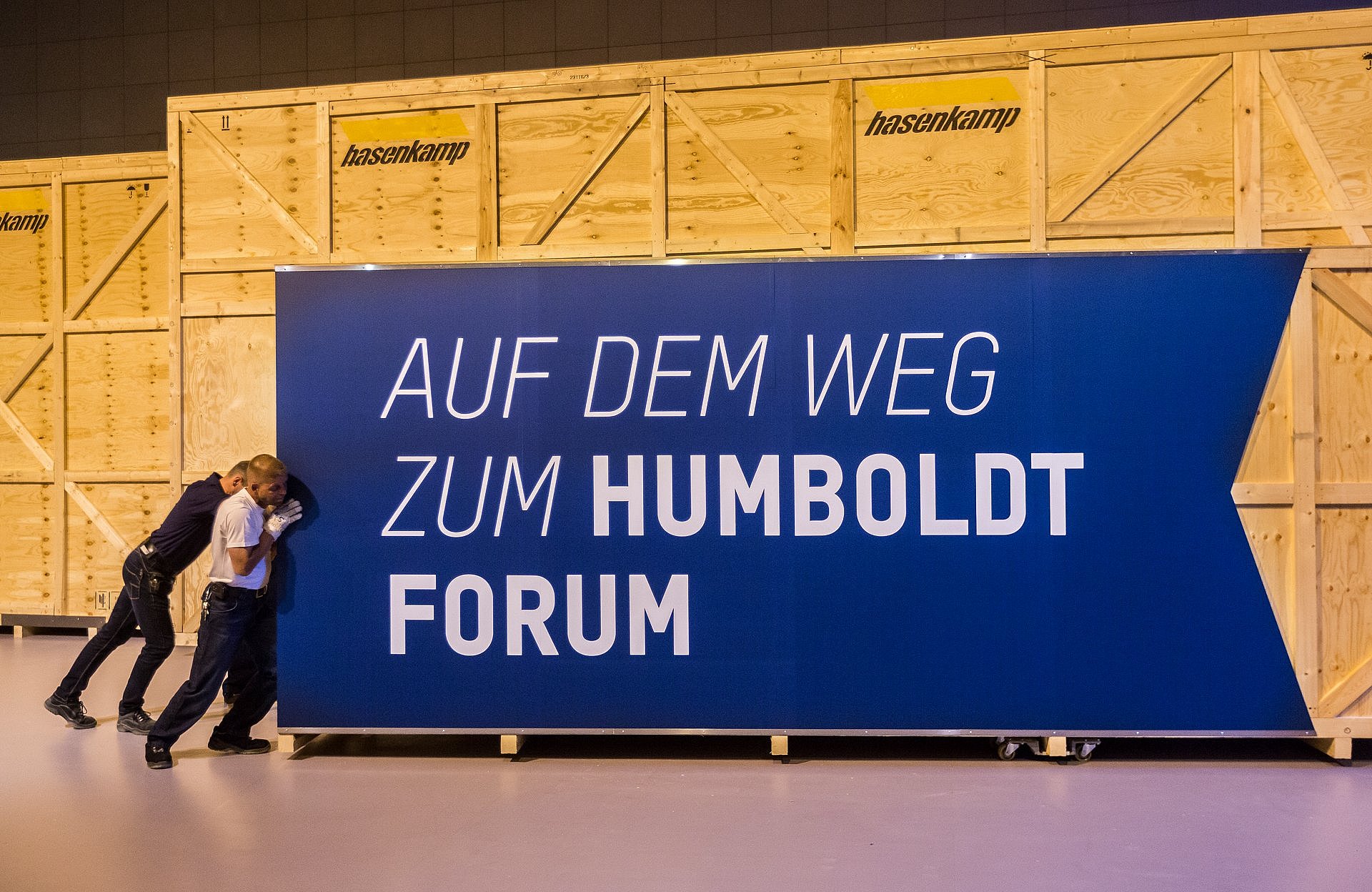 Zwei Männer schieben eine überdimensionierte Kiste, die mit dem Schriftzug "Auf dem Weg zum Humboldt Forum" beklebt ist