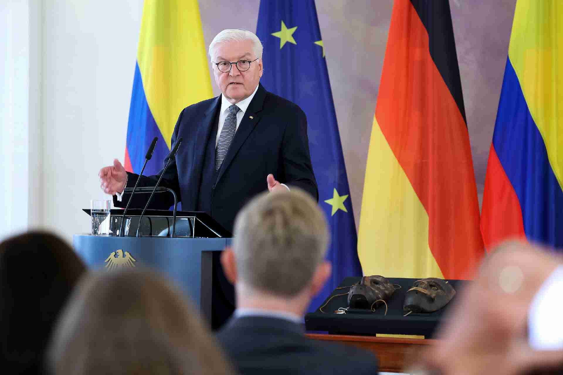 Weißhaariger Mann hält Rede vor Publikum mit Masken neben ihm