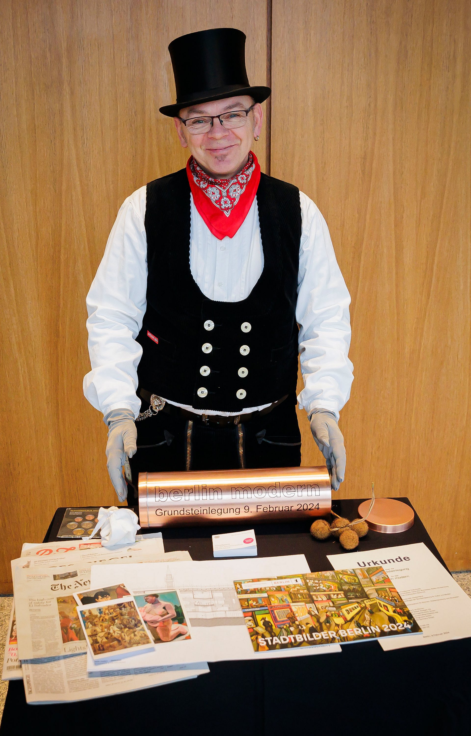 Ein Mann in Handwerkskleidung hält eine metallische Hülse, vor ihm liegen Gegenstände auf einem Tisch