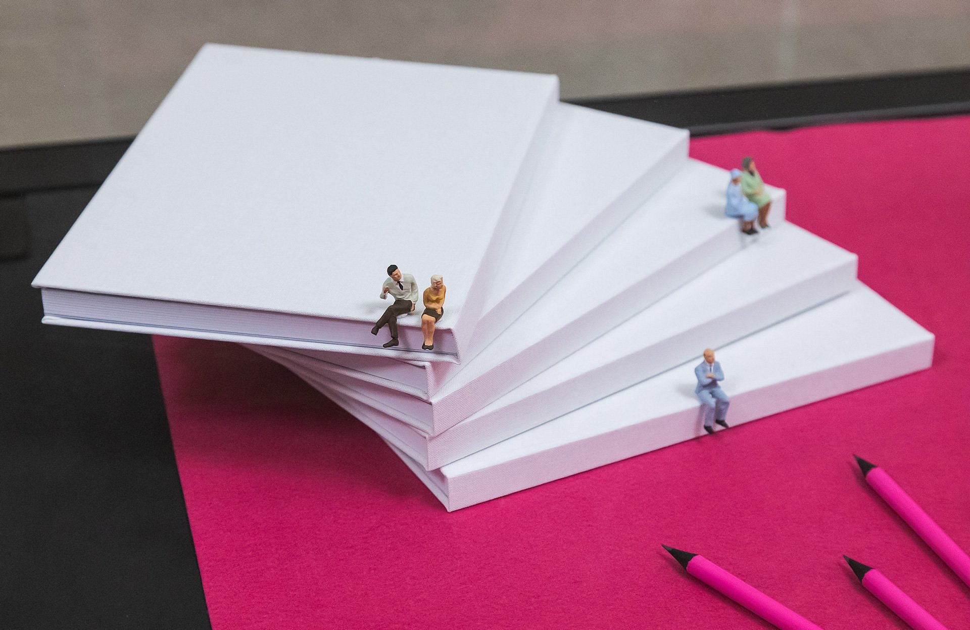 Gestapelte leere, weiße Bücher, darauf kleine Figuren, pinker Untergrund