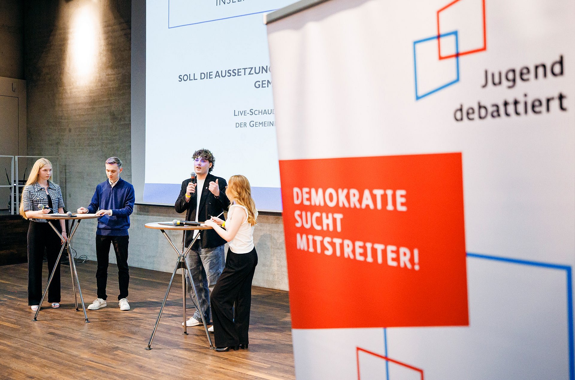 Vier Personen stehen an einem Stehtisch und diskutieren, rechts im Bild ein Aufsteller mit der Aufschrift "Jugend debattiert"