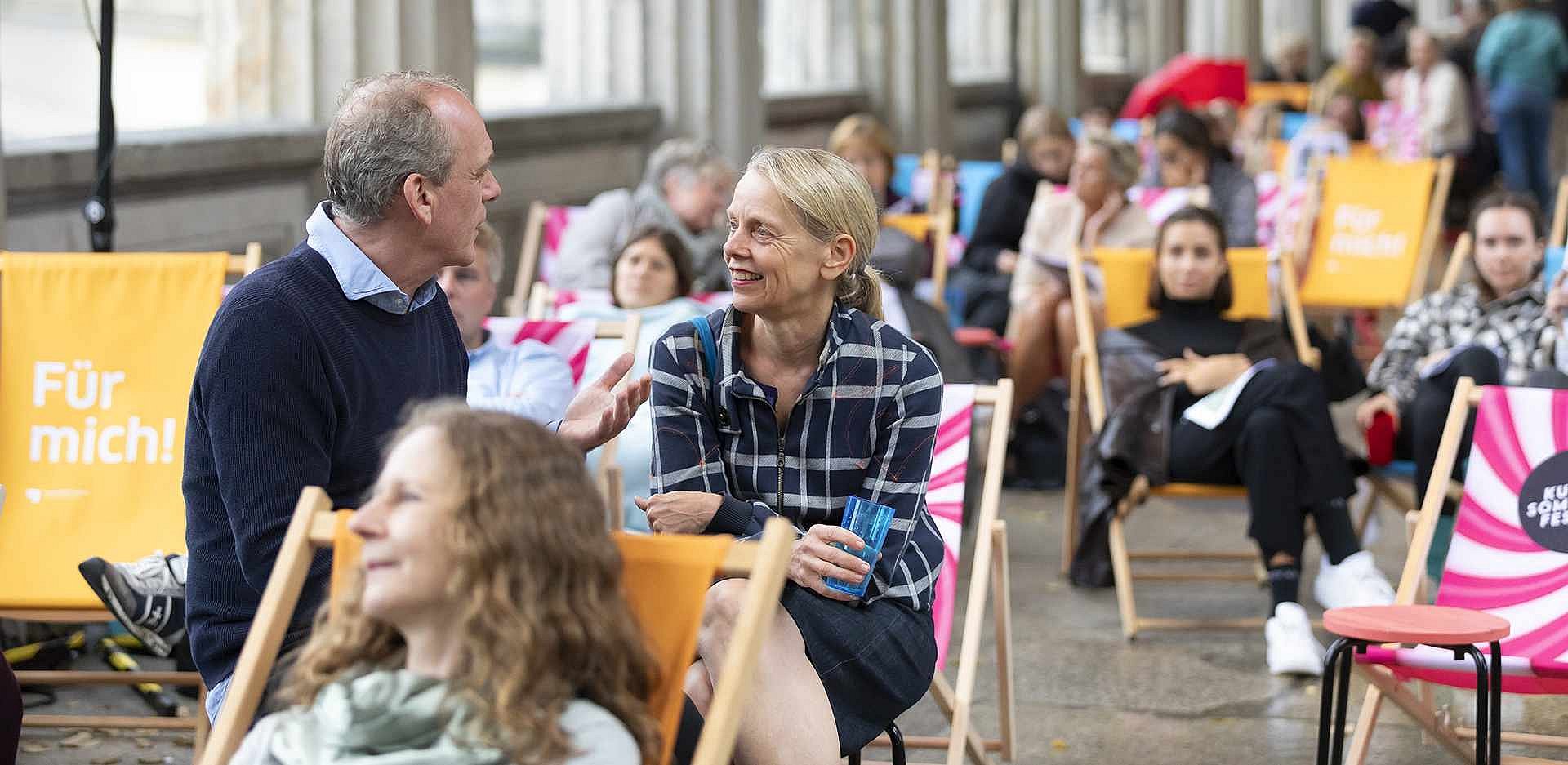 Zwei Menschen im Gespräch, auf Strandstühlen sitzend, in Menschenmenge