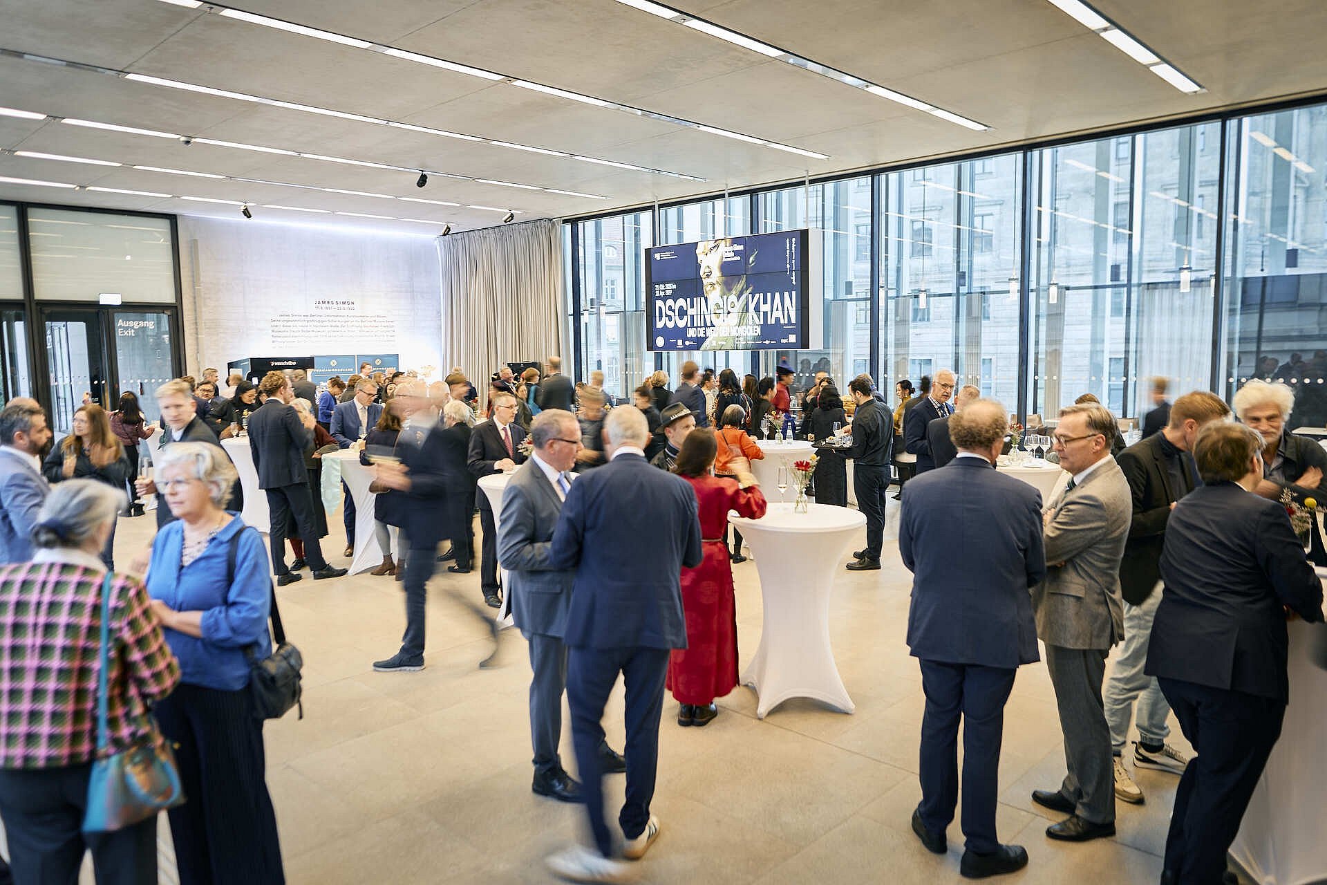 Zahlreiche Gäste unterhalten sich in kleinen Gruppen an Stehtischen im hellen Foyer. Im Hintergrund hängt das Ausstellungsplakat.