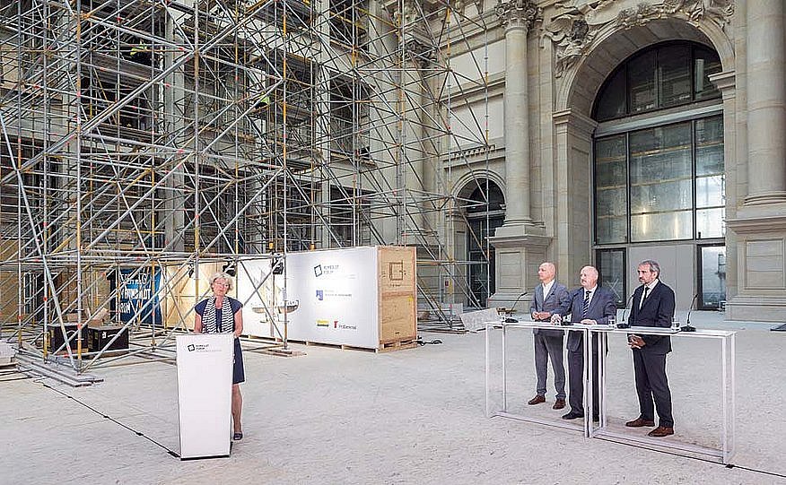 Eine Frau links im Bild spricht an einem Rednerpult, rechts stehen drei Männer an einem langen Tisch. Im Hintergrund sieht man die eingerüstete Fassade eines Gebäudes (öffnet Vergrößerung des Bildes)