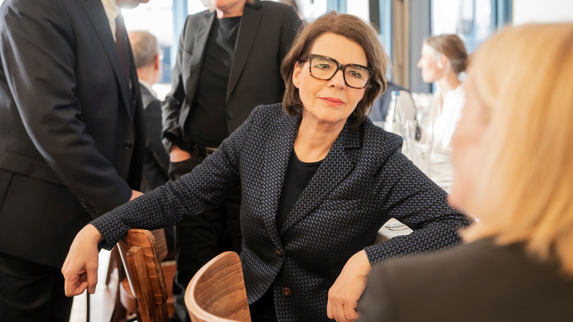 Frau mit Brille und dunklem Jackett im Gespräch bei einer Tischgesellschaft