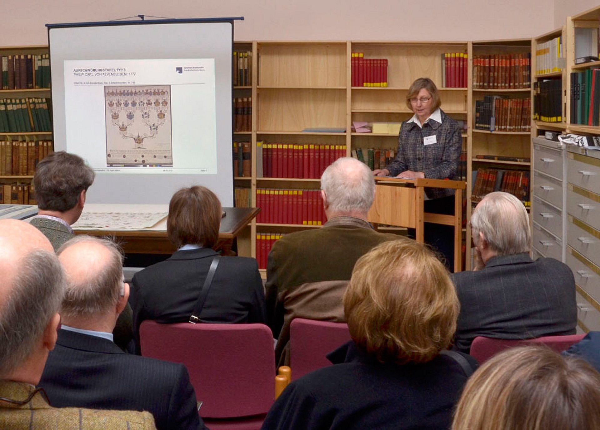 Frau spricht neben einer Leinwand zu Publikum in einer Bibliothek