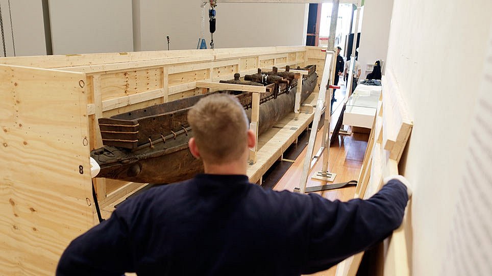 Ein Mann blickt auf eine an einer Seite offene Holzkiste, in der sich ein hölzernes Boot befindet (öffnet Vergrößerung des Bildes)