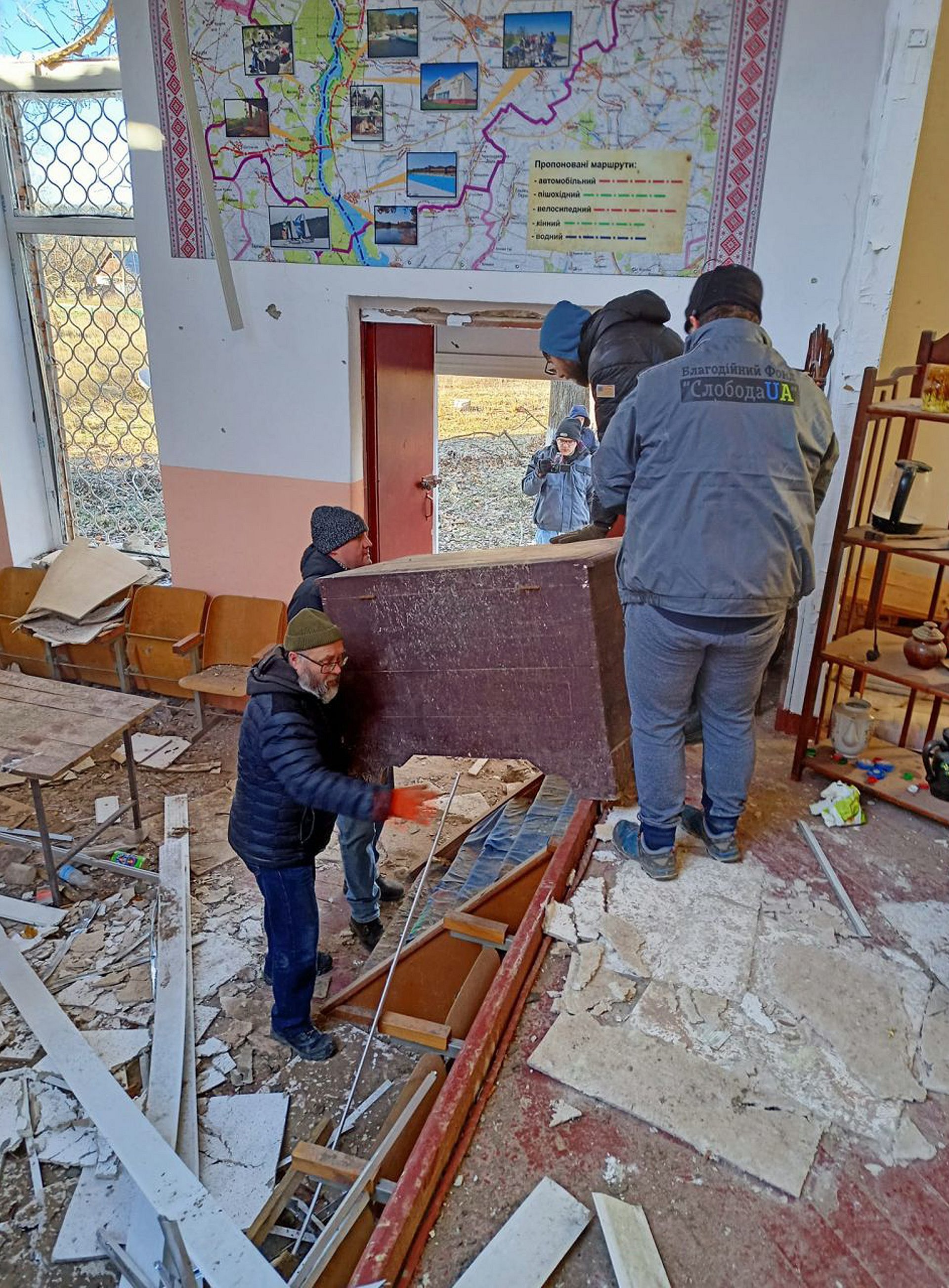 Drei Personen tragen eine Kommode durch ein beschädigtes Haus mit Trümmern auf dem Boden