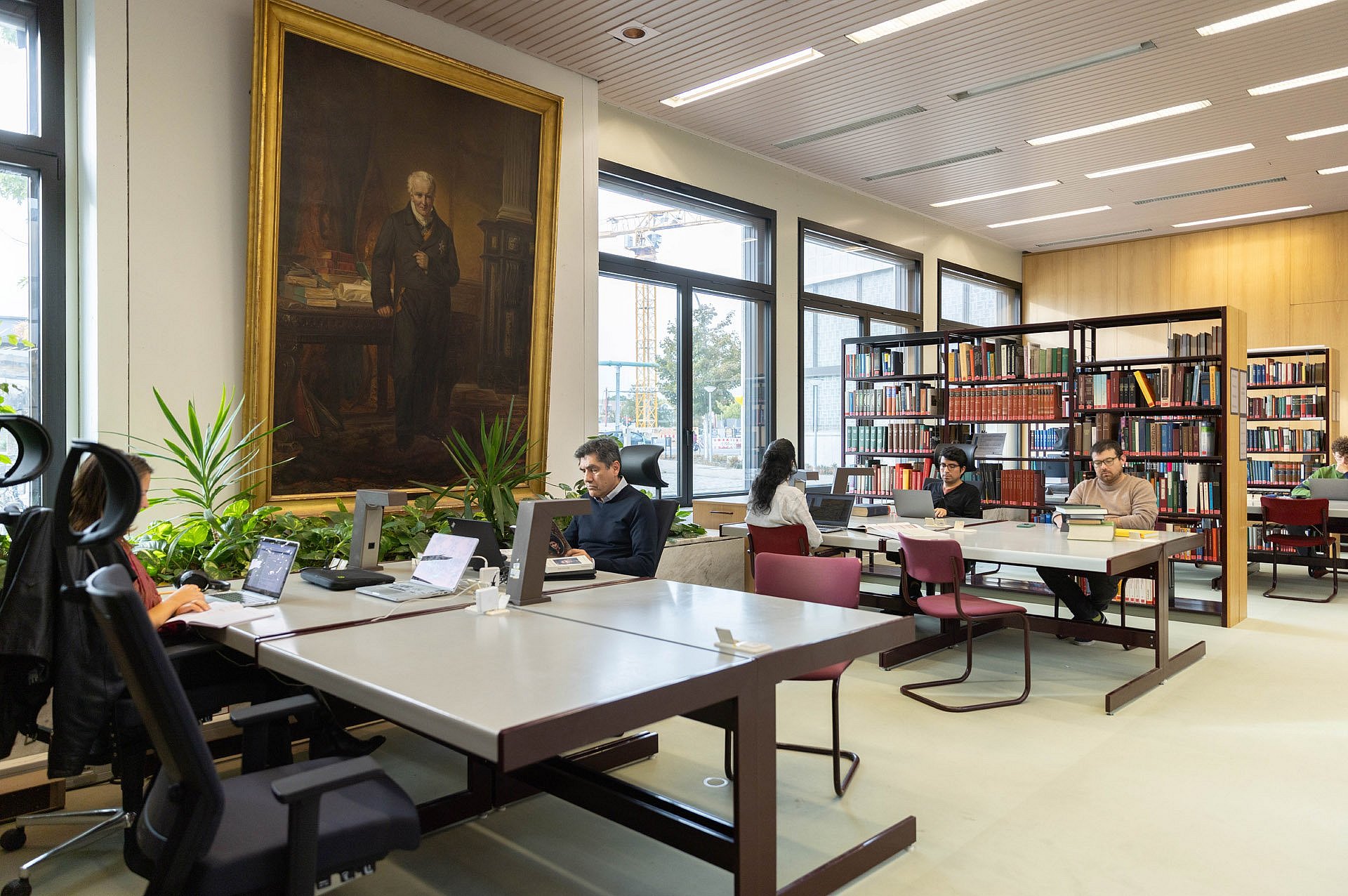 Menschen sitzen in einem Lesesaal einer Bibliothek, an der Wand hängt ein lebensgroßes Ölgemälde A.v. Humboldts