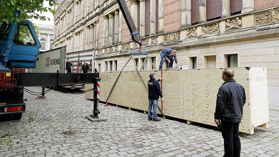 Eine große Holzkiste und mehrere Personen steht auf einem geplasterten Platz vor einem Fahrzeug (öffnet Vergrößerung des Bildes)