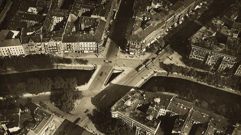Landwehrkanal, Blick von Süden mit der Doppelbrücke von Potsdamer Straße und Victoriastraße (1919): In einem der Eckhäuser an der Potsdamer Straße liegen Ernst Rowohlts Verlagsräume. In der Straße Am Karlsbad, ganz unten im Bild, hat Mies van der Rohe sein Büro (öffnet Vergrößerung des Bildes)