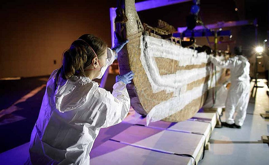 Ein Boot wird in einem musealen Innenraum von mehreren Personen verpackt und zum Transport vorbereitet. Im Vordergrund sieht man eine Frau in weißer Schutzkleidung und mit blauen Handschuhen, die das Boot stützt (öffnet Vergrößerung des Bildes)
